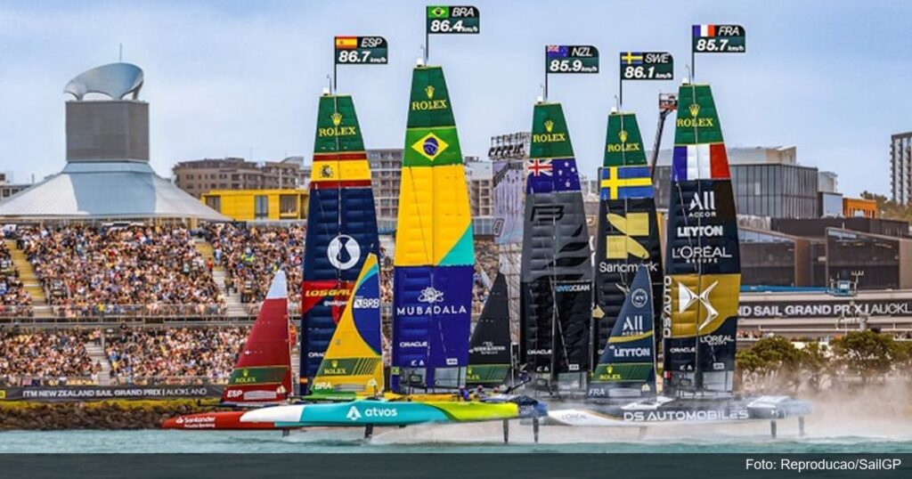 Catamarãs do SailGP disputando corrida com bandeiras de velocidade e arquibancadas lotadas ao fundo